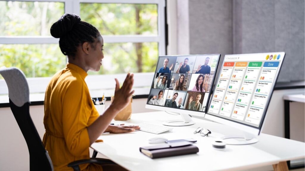 Mulher participando de uma reunião por videochamada, com uma tela de computador mostrando um quadro Kanban à sua frente, e várias pessoas visíveis em outra tela.