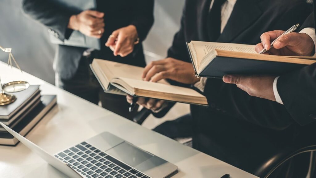Pessoas de terno reunidas em volta de um notebook trocando livros e documentos, sugerindo um ambiente jurídico.