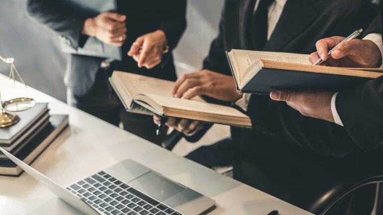 Pessoas de terno reunidas em volta de um notebook trocando livros e documentos, sugerindo um ambiente jurídico.