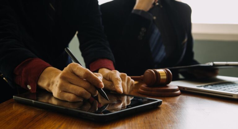 ois advogados trabalhando em um escritório, um assinando documentos em um tablet e outro usando um martelo de juiz, simbolizando a prática jurídica e o processo judicial.