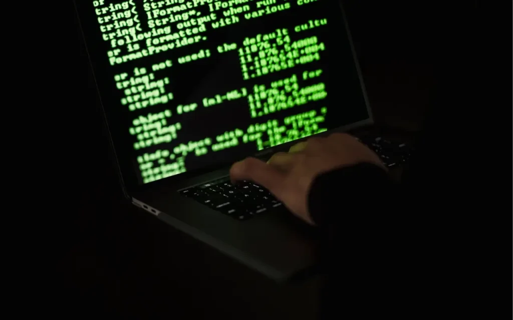 Pessoa usando um laptop com tela de código de programação verde, simbolizando hacking ou segurança digital, focado na atividade de cibersegurança.
