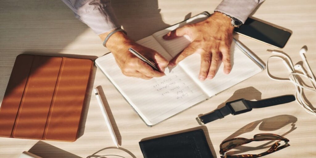 imagem de mão anotando em agenda, em mesa com utensílios de escritório