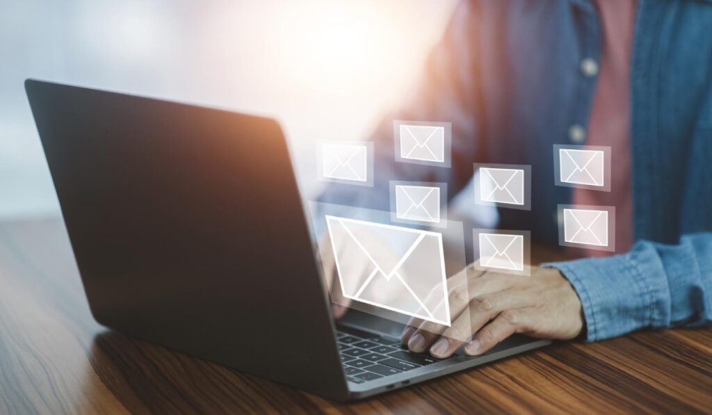 Pessoa digitando em um laptop com ícones flutuantes de envelopes de e-mail ao redor, representando comunicação eletrônica e gerenciamento de e-mails.