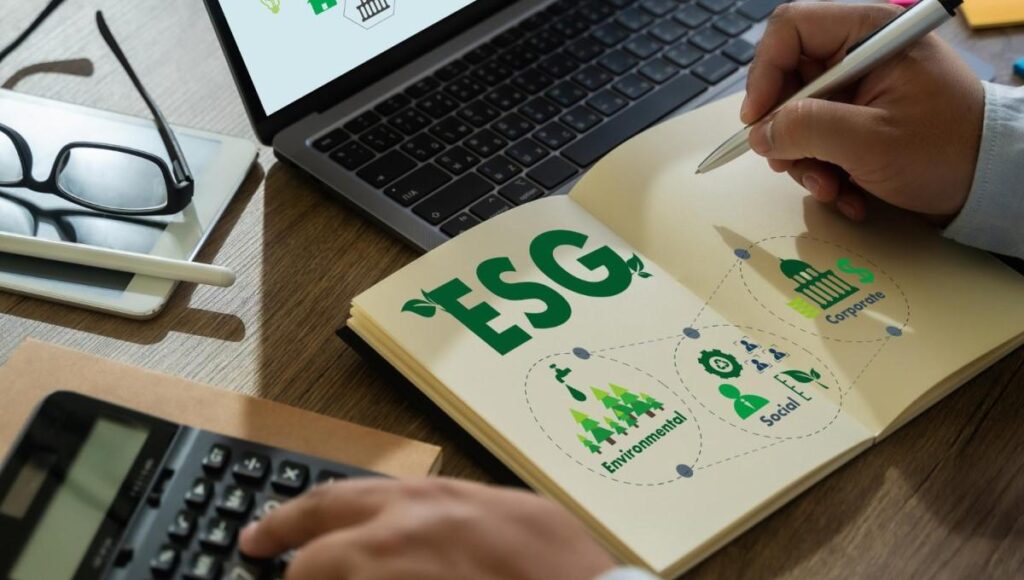 Mão segurando caneta e anotando em um caderno com gráficos e texto "ESG", ao lado de um laptop, calculadora e óculos, simbolizando a análise de práticas empresariais sustentáveis.