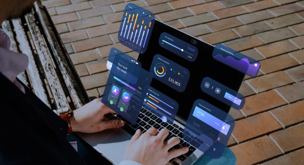 
Pessoa utilizando um laptop ao ar livre, com gráficos e dados digitais flutuando na tela. A imagem mostra várias janelas virtuais exibindo diferentes tipos de gráficos, indicadores e informações analíticas. O cenário ao fundo sugere um ambiente urbano com pavimentação de tijolos. A pessoa parece estar analisando dados ou monitorando informações em tempo real, combinando trabalho digital com mobilidade.