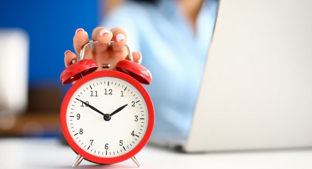 Pessoa segurando um despertador vermelho em frente a um laptop, simbolizando gestão de tempo.