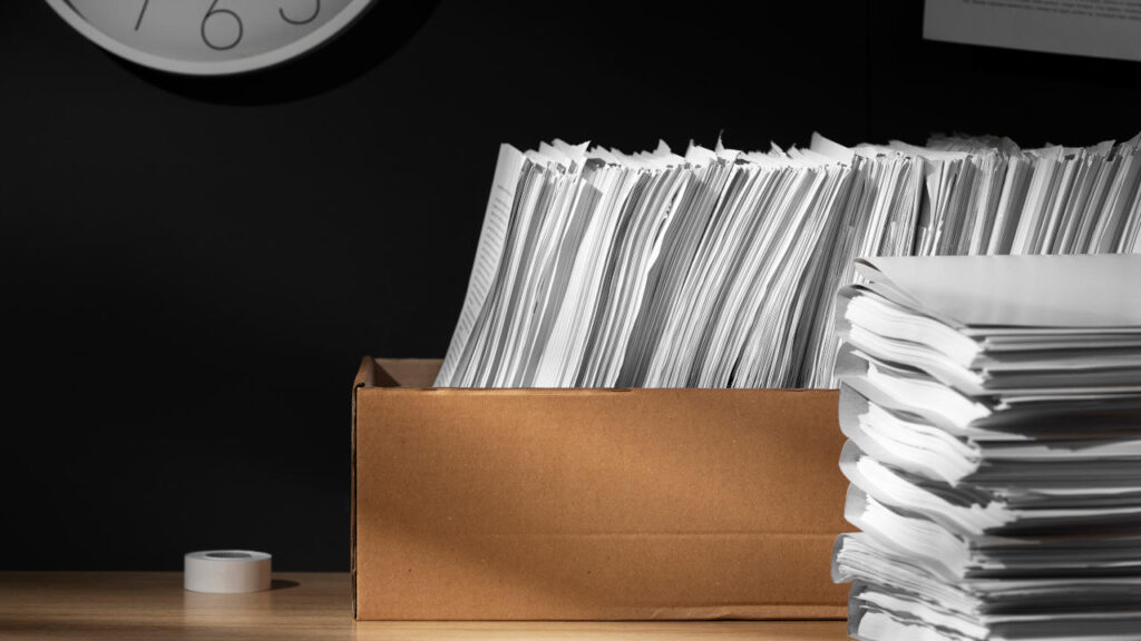 Pilha de documentos em uma caixa de papelão, sugerindo grande volume de papel em um ambiente de trabalho.