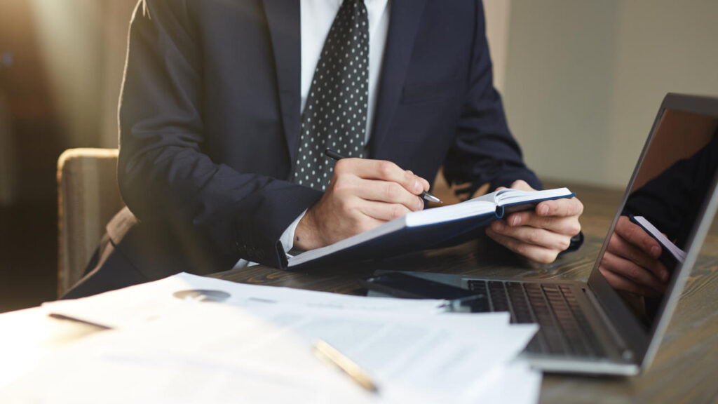 Pessoa em traje formal fazendo anotações em um caderno em uma mesa de escritório ao lado de um laptop.
