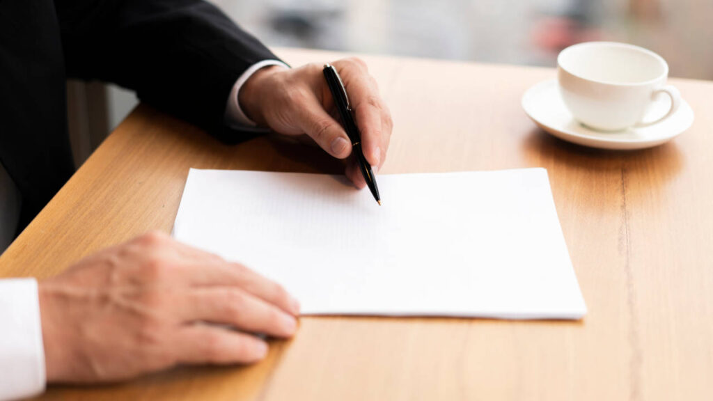 Pessoa assinando um documento em uma mesa com uma xícara de café ao lado.