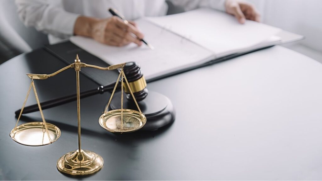 Homem escrevendo em um papel, com um martelo de advogado e o símbolo da balança da justiça visíveis na mesa ao seu lado.