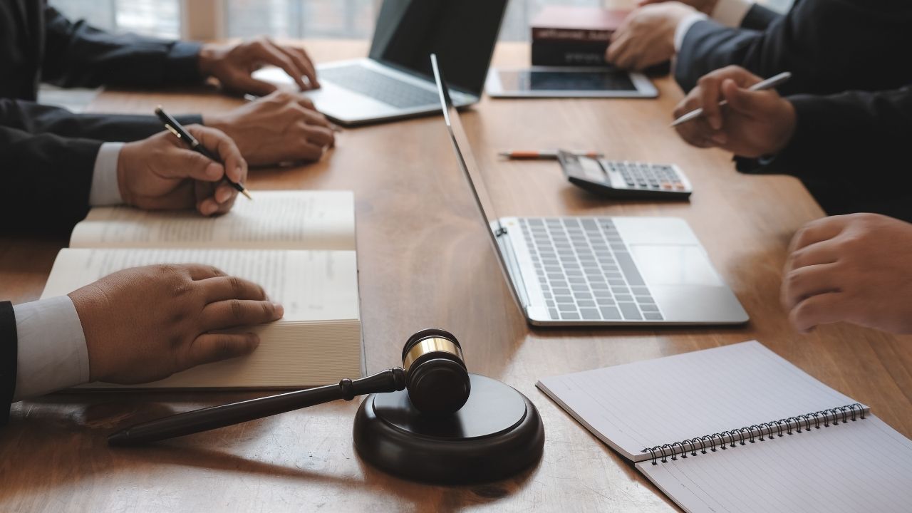 Homem digitando em um notebook, com outros notebooks e papéis espalhados sobre a mesa, enquanto um martelo de advogado está ao lado.