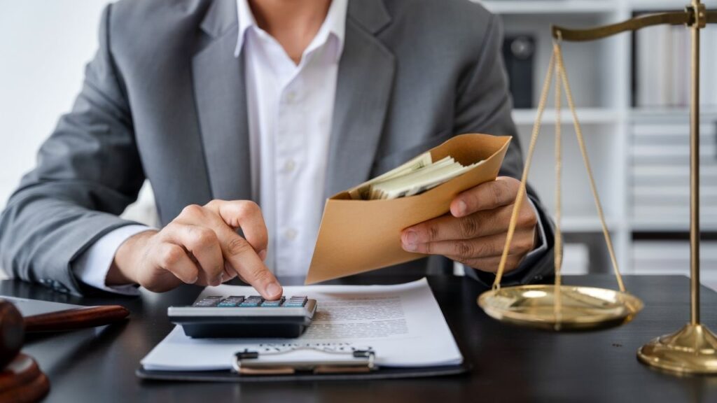 Um homem de terno segurando um envelope ao lado de uma balança da justiça, enquanto usa uma calculadora.