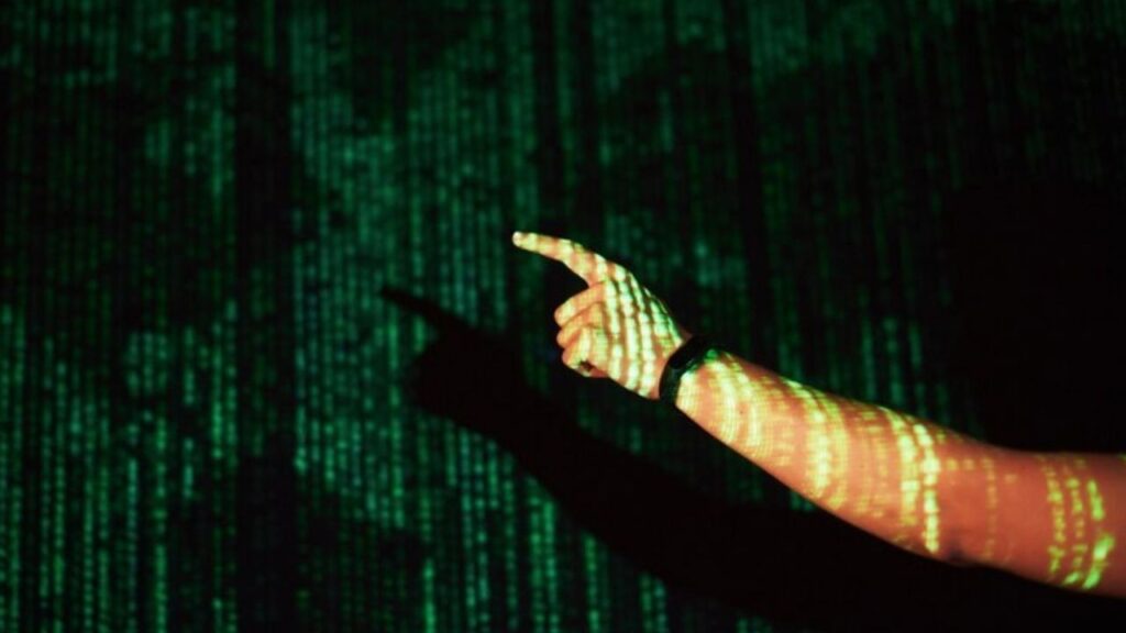 Imagem que mostra uma mão apontando para a frente, com padrões de código em verde refletidos na pele. Representa temas como tecnologia e programação.