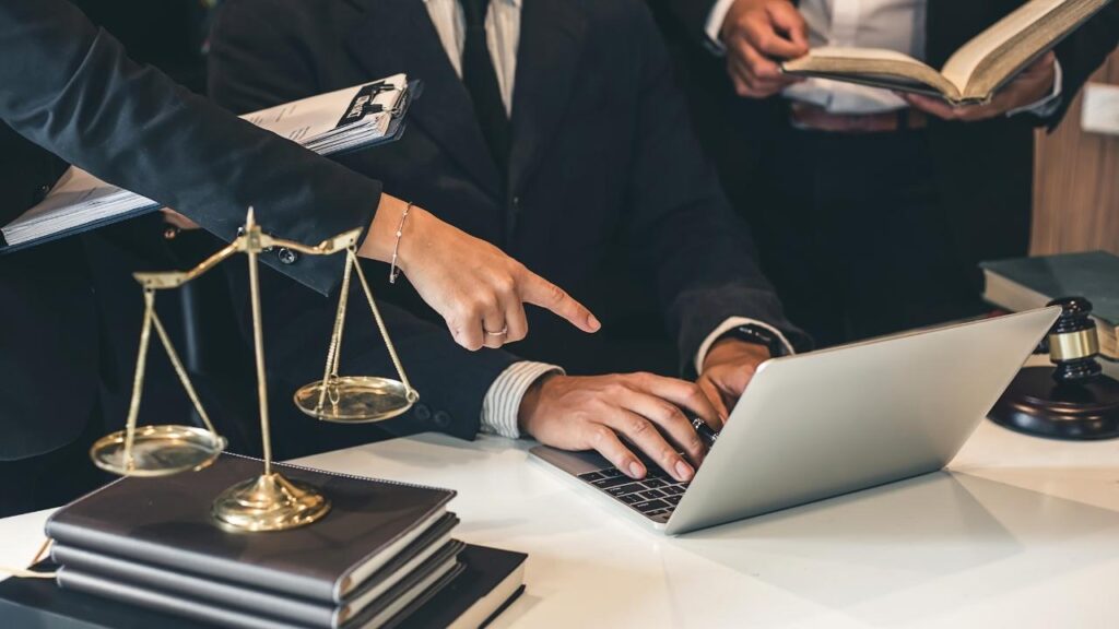 Mãos de uma pessoa digitando no notebook, com outra mão apontando para a tela, e uma balança dourada ao lado, representando um trabalho judiciário.
