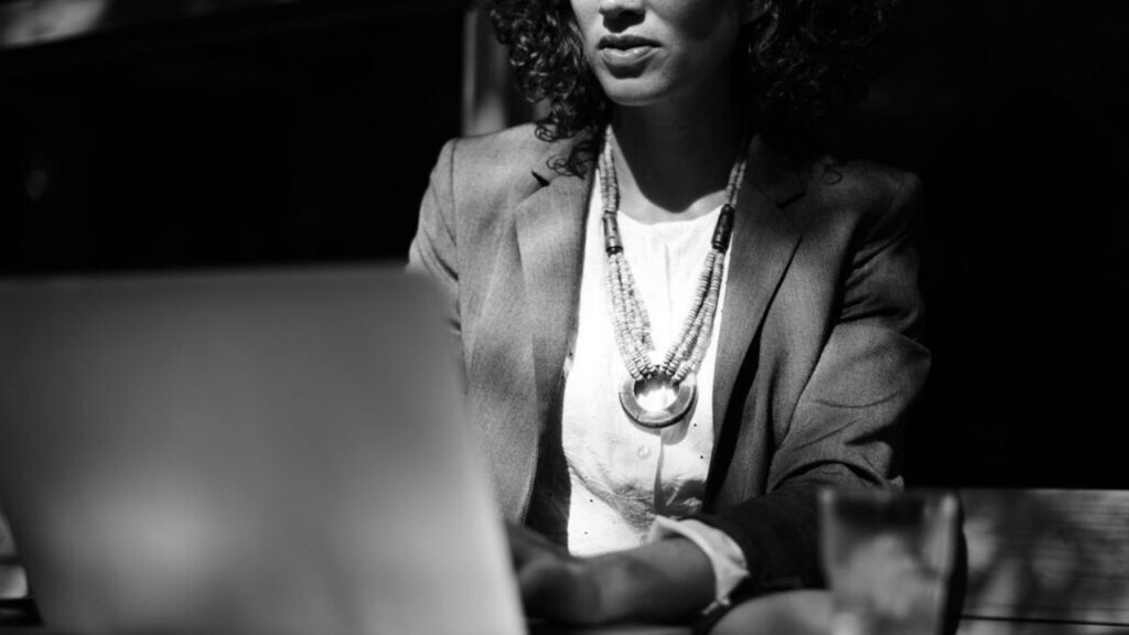 Mulher de negócios sentada, usando um blazer e colar, concentrada no laptop em um ambiente iluminado. A imagem destaca a formalidade e a produtividade.