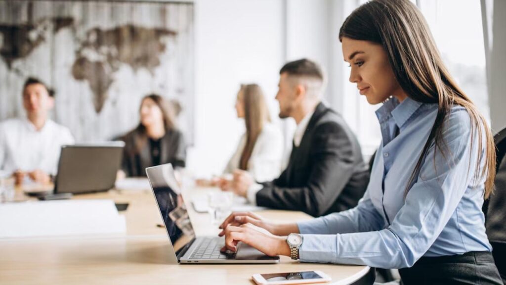 Mulheres e homens em uma reunião de negócios, debatendo ideias e utilizando laptops em uma mesa de trabalho moderna.