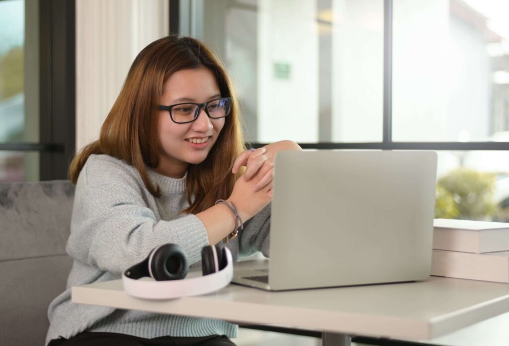 Uma jovem adolescente asiática sorridente, com cabelos castanhos claros e óculos pretos, usa um suéter cinza e interage com um laptop prateado à sua frente. Fones de ouvido brancos e pretos estão na mesa ao lado do laptop, e alguns livros brancos estão empilhados no lado direito. Ela está sentada em um sofá cinza em um ambiente bem iluminado, possivelmente um café ou uma biblioteca, com grandes janelas ao fundo que mostram um dia claro.