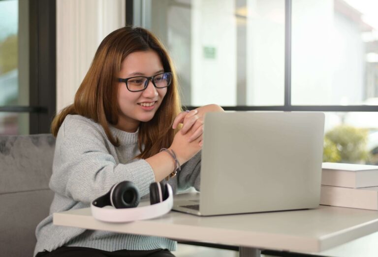 Uma jovem adolescente asiática sorridente, com cabelos castanhos claros e óculos pretos, usa um suéter cinza e interage com um laptop prateado à sua frente. Fones de ouvido brancos e pretos estão na mesa ao lado do laptop, e alguns livros brancos estão empilhados no lado direito. Ela está sentada em um sofá cinza em um ambiente bem iluminado, possivelmente um café ou uma biblioteca, com grandes janelas ao fundo que mostram um dia claro.